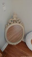 Full front view of the ornate oval mirror showing cream-colored detailed frame with floral and urn motifs and oval glass.
