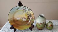 Porcelain plate with a painted scene of goats near birch trees along with two painted porcelain vases on a lace tablecloth.