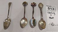 Four sterling silver spoons displayed side by side on white background, two with colorful crest emblems, two plain spoons