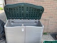 Open storage box showing interior and hinged green lid raised against brick wall