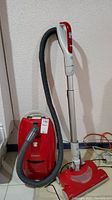 Full view of the vacuum cleaner standing upright showing the red main unit, grey hose attached to the wand and floor nozzle with red and white handle.