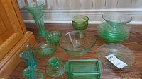 Overall view of vintage green glass dishes on wooden floor including bowls, plates, candle holders, butter dish, vase, creamer, and ashtray.