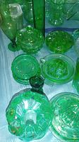 Photo shows various vintage uranium green glass items including vases, candy dish with lid, flower frog, juicer, bowls, and plates on a table.