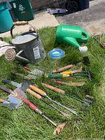 Overview of all items laid on grass: metal watering can, plastic watering can, gardening tools such as trowels, weed pullers, hand rake, and coil of blue wire.
