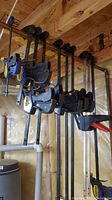 Wide view showing six Irwin Quick-Grip bar clamps hanging from ceiling beam in basement area with wood and pipe background.