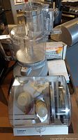 Photo showing Cuisinart food processor with clear plastic bowl and feed tube, and the blade/disc holder in front.