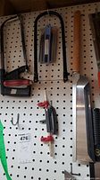 Various saws, including hacksaws with red and black handles, hand saws, and saw blade replacements hanging on pegboard