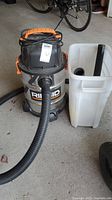 Ridgid stainless steel shop vacuum with black flexible hose and cord wrapped on top, situated next to a white plastic container holding additional vacuum accessories.