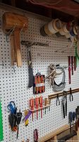 Overall view of tools hanging on white pegboard with wooden mallets, hammer, screwdrivers, scissors, and vice grips.