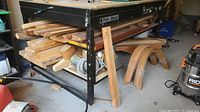 Photo showing hardwood lumber stacked under a metal table, includes straight planks of varying sizes and bent wood pieces on the floor wrapped in tape.