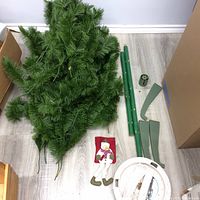 View of disassembled artificial Christmas tree sections including green branches and tree poles, along with two metal stands and a snowman stocking on wooden floor near cardboard boxes