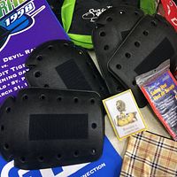 Three black knee pads with vent holes laid flat, accompanied by other items partially visible such as safety vest packaging and boxed small items.