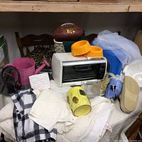 Shows collection of household items on table including Black & Decker toaster oven, football, Crocs shoes, watering can, dog coat, ceramic mugs, and laundry bags.