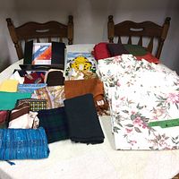 Photo of assorted ladies scarves and folded curtain panels displayed on a table and chairs in background.