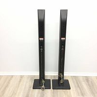 Rear view of two slim black LG tower speakers on square bases showing speaker wire connections and repaired stand with black caulking on one stand.