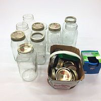 Six clear glass Mason jars of varying shapes and sizes arranged upright. Several metal lids on top of some jars. Next to the jars is a brown cardboard box filled with numerous metal lids and bands. There is a blue box with additional lids on the side.