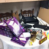Photo showing 9 unopened boxes of Avery cord-style name badges along with various other office supplies such as black desk trays, calculator, clipboards, brass jacket organizer, and rolls of paper on a table.