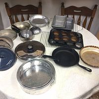Photo of entire collection of pots, pans, bakeware and kitchen items arranged on a table, showing variety including cast iron skillet, glass lid pot, muffin tins, cooling racks, pie pan.