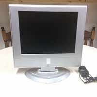 Front view of the Prima 15 inch LCD TV with silver frame, black screen, and silver round stand. Power adapter placed to the side.