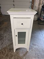 Front view of the small white bathroom cabinet showing the drawer and fogged glass door.
