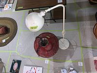 White tall floor lamp with curved neck and round lamp head upright next to the red wicker hanging lamp shade on the ground.