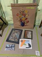 Five art pieces displayed on the floor and the needlepoint tapestry hanging on a rod against the wall.