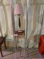 Photo of tall metal plant stand with ornate circular design on top, supporting teak table lamp with white fabric shade.