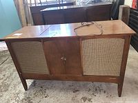 Front view of the wooden record player cabinet showing the woven speaker grills, central cabinet door with handle, and polished wood finish.