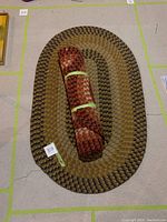 Photo of two oval braided rugs; one large laid flat in brown, beige, and black colors, and one smaller rolled-up rug in red and orange tones placed on top.
