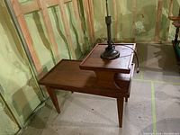 Two tier wooden side table with tapered legs and stepped design, photographed in unfinished room.