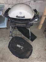 Front view of white Weber tabletop BBQ with lid closed, placed on a wheeled metal stand, and black Weber cover on the floor in front.
