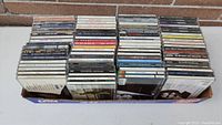 Large variety of approximately 80 CDs stacked in rows inside a box, view showing spines and top edges of jewel cases
