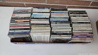 Wide view of about 85 CDs stacked in a box showing spines with artist and album titles, cases dusty and needing cleaning.