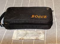 Closed black soft bocce ball carrying case with 'BOCCE' label and a visible printed information sheet beneath it on a fabric surface.