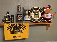 Wide shot showing all items displayed on wooden shelf against wall: Bruins sign, plush moose, bear logo towel, hockey player figurine, koozie, Tim Hortons card, small flag and hockey puck.