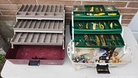 Two fishing tackle boxes with multiple compartments, one maroon-colored and the other green, filled with assorted fishing lures and hooks. Photos show the overall lot setup and condition.