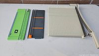 Three paper cutters laid out on a white surface: green Cricut scoring board on left, black X-Acto rotary trimmer in middle, beige guillotine cutter on right.