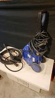 Blue electric pressure washer unit with power cord wrapped on handle, showing two large rear wheels and vented sides.