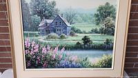 Frontal view of framed oil painting depicting farmhouse by pond with pink and white flowers, green trees and distant hills