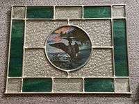Full view of stained glass window hanging showing rectangular design with textured clear and green glass panels surrounding a central circular panel featuring a painted loon on water.