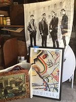 Framed Kandinsky print alongside large Beatles poster, items shown leaning against a chair and wall in storage area.