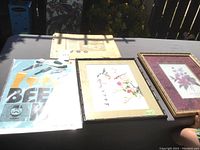Four artworks displayed on a table including Beer Me poster, embroidered piece, floral print, and a squeegee plate