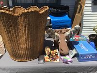 Wide shot displaying tall woven wicker basket, fabric moose head, board games, wooden duck figurine, comic books, and small box on outdoor table surface.