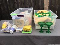 Two large clear plastic bins filled with assorted paper products including napkins and disposable cups along with two green glass margarita-style glasses placed in front.
