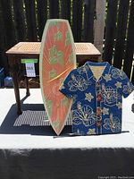 Full lot view showing surfboard clock, Hawaiian shirt corkboard, and woven seat stool on table outside.
