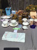 Photo of assorted teacups, saucers, votive candle holders, Japanese teapot set, and embroidered cloth laid out on a table