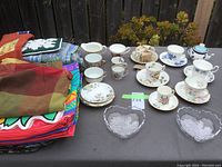 Overview of assortment of teacups, saucers, glass dishes, and folded table linens displayed on outdoor table.