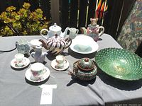 Wide view of assortment of teapots, teacups, saucers, and decorative bowl on table outside.