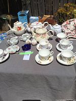 Full view of assortment of teapots, teacups, saucers, creamer, and decorative vases arranged on table covered with gray cloth, outdoor lighting.