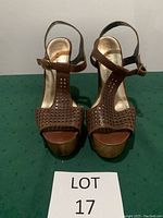 Front view of brown Baker platform sandals with studded front strap and ankle buckle closure, displayed on green surface with LOT 17 label.